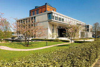 Stiftungsgebäude in Hamburg-Langenhorn. Blick auf das Gebäude und die grüne Hecke drum herum. Es ist eine moderne Architektur, ebenfalls ist der alte Kaffeeturm zu sehen, um den sich das neue Gebäude herumzieht.